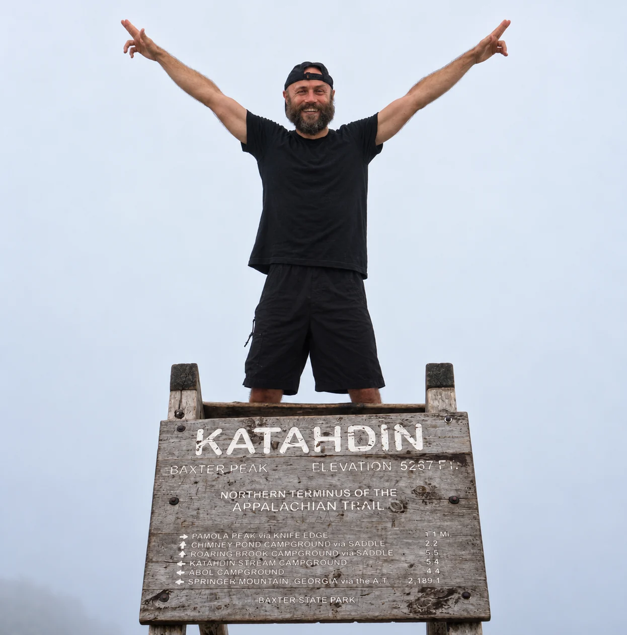 Justin Bazan with both arms raised behind the Katahdin summit sign