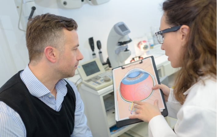 An optometrist seated down with her male patient using a detailed photo of an eye to explaining the effects of glaucoma