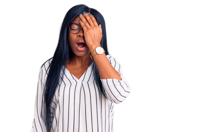 A woman wearing casual clothes in front of a white background holding her eye with her hand and mouth open as she groans in pain