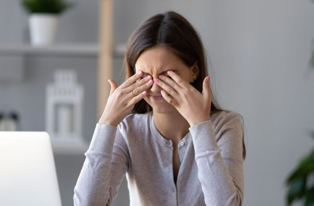 A lady in a gray colored cardigan is touching her closed eyes with a gesture of irritation on her face.