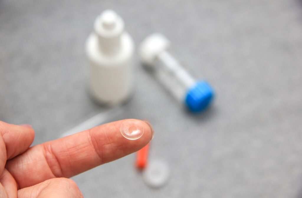 A close-up of a person's hand, with a contact lens resting on their fingertip. In the background, a contact lens case and a bottle of cleaning solution sit on a table.